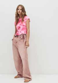 Růžový tie-dye top s krátkým rukávem v kombinaci s volnými, světle růžovými džíny s elastickým pasem a bočními kapsami. Sandály se viditelnými popruhy.