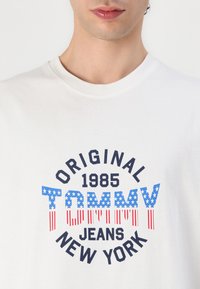 White cotton t-shirt features a circular blue, red, and black graphic with "TOMMY" and decorative star and stripe elements.