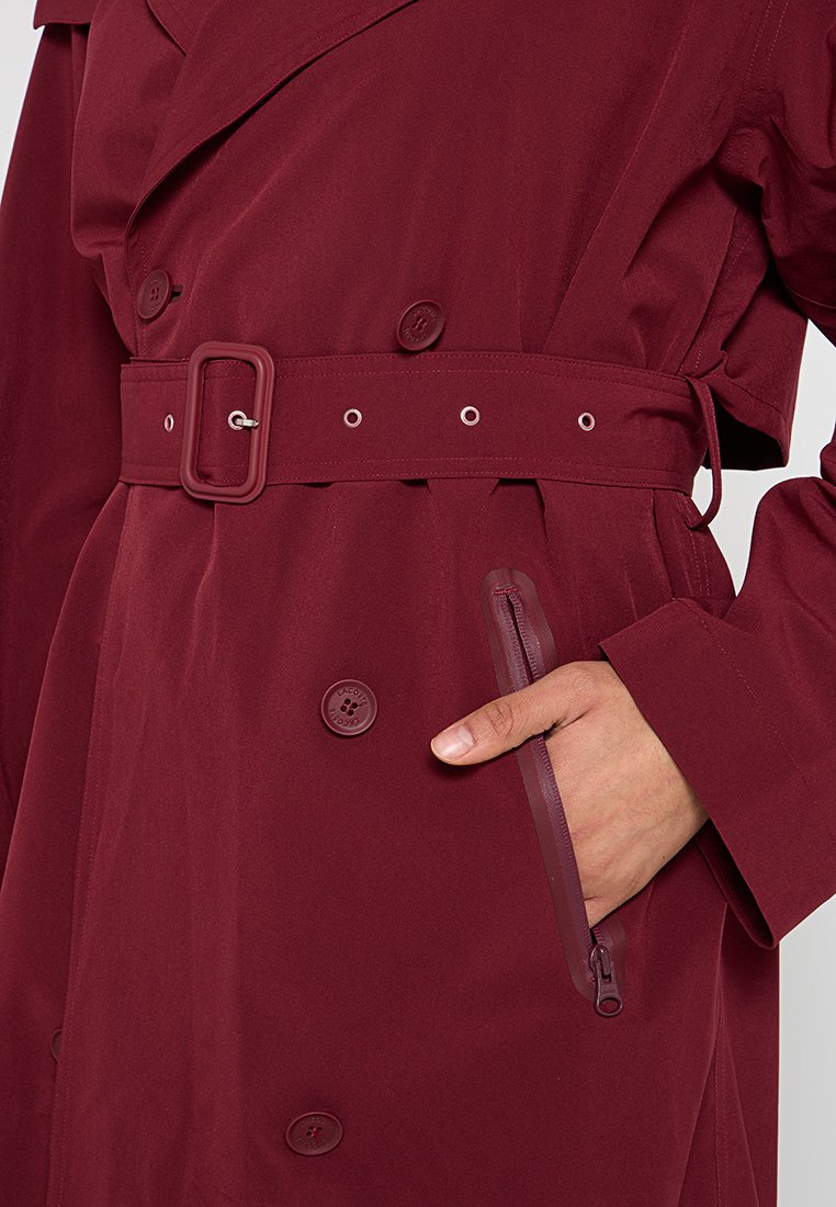 Bordeauxkleurige trenchcoat met een ceintuur in de taille, dubbelzijdige knopen en een ritssluiting zak. De stof lijkt glad en waterafstotend te zijn.