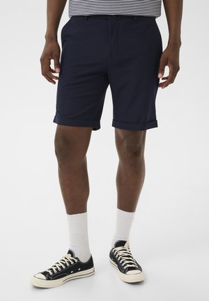 Férfi tengerészkék felhajtott rövidnadrágban, fehér, a lábszár közepéig érő zokniban, fekete alacsony szárú tornacipőben és fekete-fehér csíkos ingben.