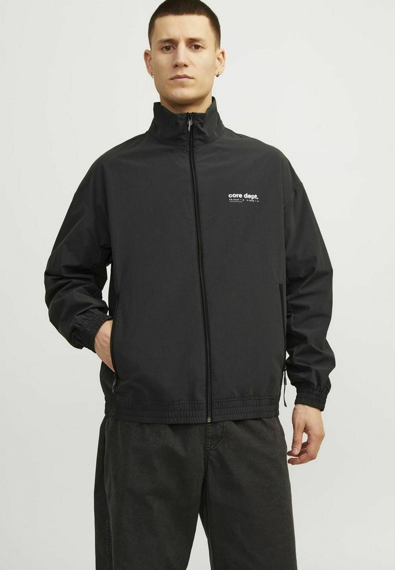 Juodas zip-up tipo švarkas su stovinčia apykakle, elastingomis rankovėmis ir juosmeniu. Kairiajame krūtinės krašte yra baltas logotipas. Lygus, lengvas audinys.