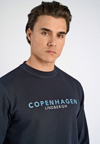 Marinefarbenes Sweatshirt mit Rundhalsausschnitt, das "KOPENHAGEN" und "LINDBERGH" in kontrastierendem Blau und Weiß auf der Brust aufgedruckt hat.
