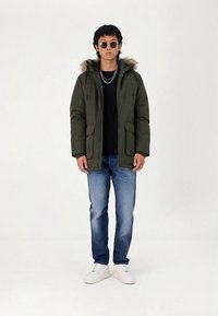 Alyvuogių žalumo parko striukė su dirbtinio kailio gobtuvu, priekine kišenėle, užtrauktuku, dėvima ant juodo marškinėlio ir mėlynų džinsų, derinama su baltais sportiniais bateliais.