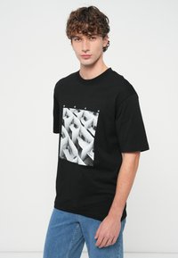 Νεαρός άνδρας με σγουρά μαλλιά, ντυμένος με μαύρο T-shirt που έχει ασπρόμαυρο σχέδιο γραφείου με κυβάκια και μπλε τζιν, σε λευκό φόντο.