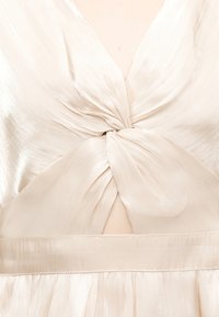 Κοντινό πλάνο εκρού σατέν φόρεμα με σχέδιο διπλωμένου κόμπου στο στήθος και ελαφρύ άνοιγμα στη μέση κάτω από το μπούστο.