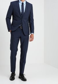 Completo blu navy con vestibilità su misura, giacca a bottone singolo e pantaloni a gamba dritta. Abbinato a una camicia azzurro chiaro e una cravatta scura.