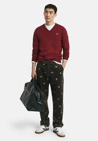 Burgunderfarbener V-Ausschnitt-Pullover, darunter ein weißes T-Shirt, dunkle Cordhosen mit Vogelmustern, schwarze Ledertasche und weiße Sneakers mit Akzenten.