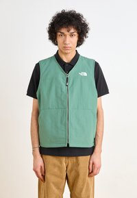 Green, sleeveless vest with front zipper, two side pockets, and a logo on the upper left. Worn over a black shirt and corduroy pants.