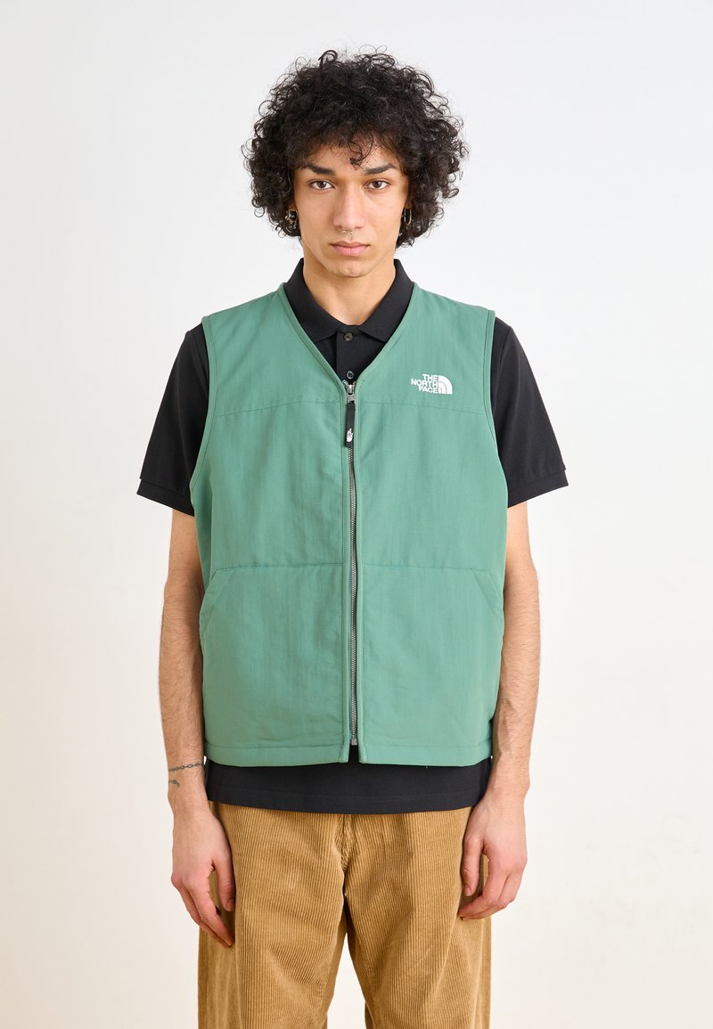 Green, sleeveless vest with front zipper, two side pockets, and a logo on the upper left. Worn over a black shirt and corduroy pants.