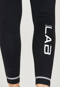 Leggings de sport noirs ajustés, ornés de l'inscription « ELITE LAB » en lettres blanches et de rayures grises aux poignets.
