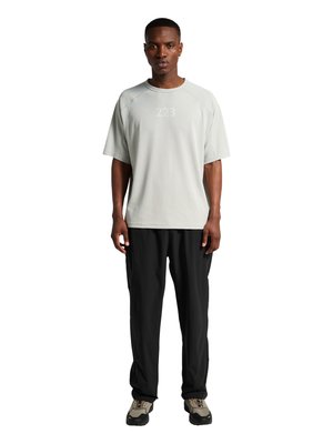 Mand iført en lysegrå kortærmet skjorte med teksten "223", sorte bukser og beige sorte sneakers, står foran på hvid baggrund.