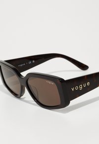 Juodos tortoiseshell saulės akinių su storu rėmeliu ir rudos spalvos lęšiais. Šone yra auksinė "vogue" logotipo detalė. Lygi tekstūra, stačiakampio formos.