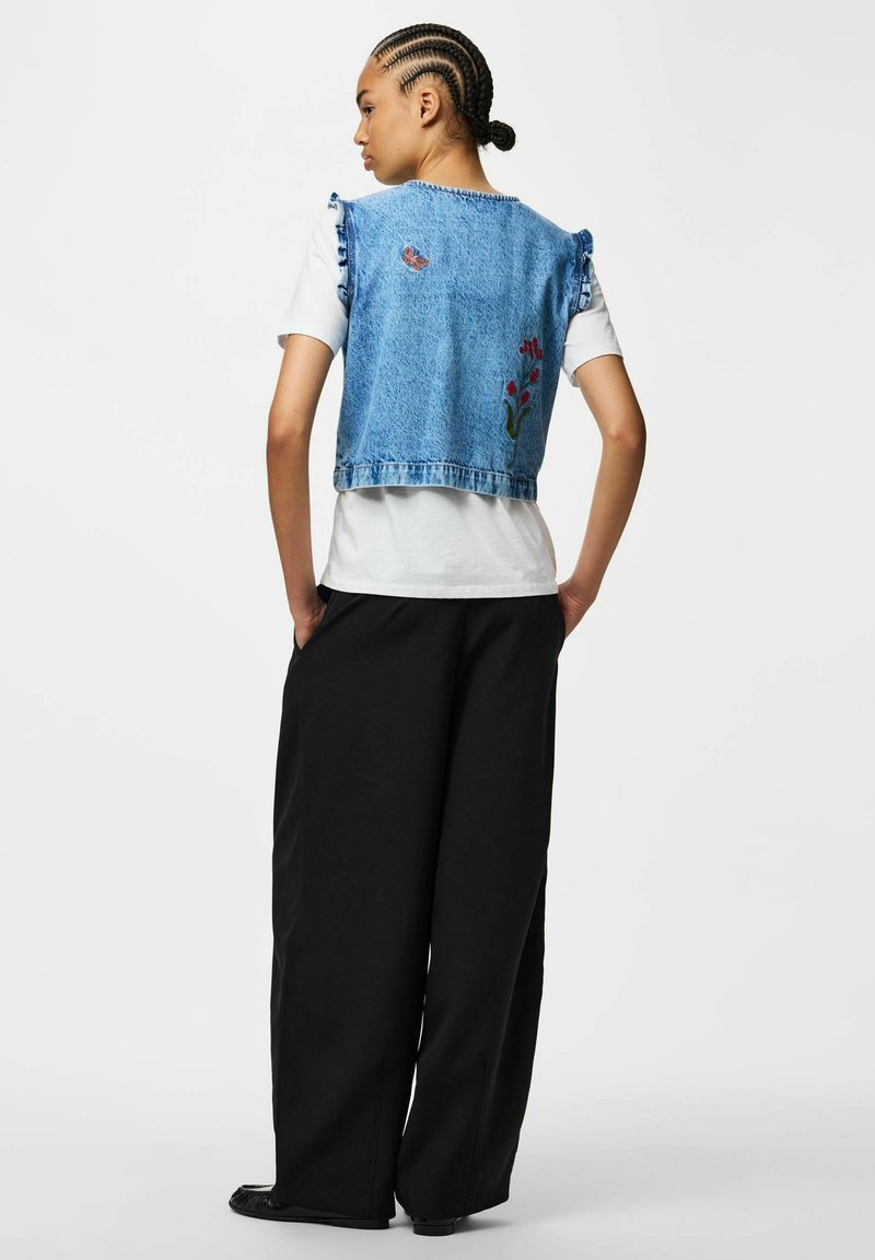 Personne aux cheveux tressés de dos, portant un gilet en jean à broderie florale, un t-shirt blanc et un pantalon noir ample, les mains dans les poches.