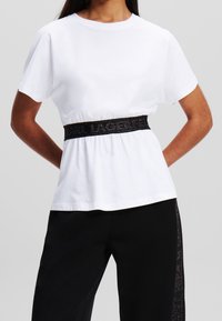 Bílé bavlněné tričko s krátkými rukávy, s černým elastickým páskem s logem. Spárované s černými kalhotami. Jednoduchý, casual design.