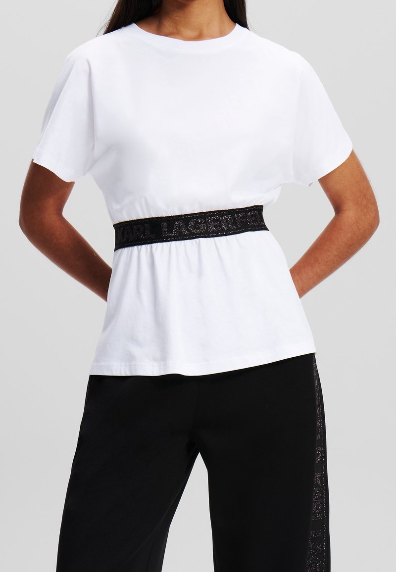 Bílé bavlněné tričko s krátkými rukávy, s černým elastickým páskem s logem. Spárované s černými kalhotami. Jednoduchý, casual design.