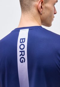 Άνδρας που φοράει μπλε πουκάμισο με κάθετη λευκή ρίγα και τη λέξη "BORG" τυπωμένη στην πλάτη, ορατός από το πλάι και πίσω.