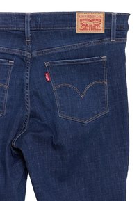 Jeans de mezclilla azul con un diseño clásico, bolsillos traseros con costura arcuada y una etiqueta de cuero marrón en la cinturilla. Detalle de etiqueta roja presente.