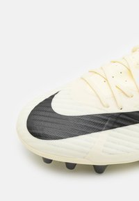 Zapatilla de fútbol Nike en color crema claro con parte superior texturizada, distintivo logo Swoosh negro y tacos negros en la suela para tracción.