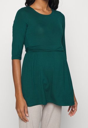 Kobieta ubrana w ciemnozielony, długi rękaw top ciążowy z marszczonym pasem, w połączeniu z jasnobeżowymi spodniami, stojąca na jednolitym tle.