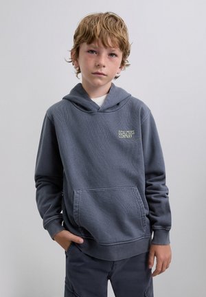 Sudadera gris con un bolsillo delantero, tela texturizada y un logotipo en el pecho. El modelo lleva pantalones oscuros a juego y tiene el cabello corto y despeinado.