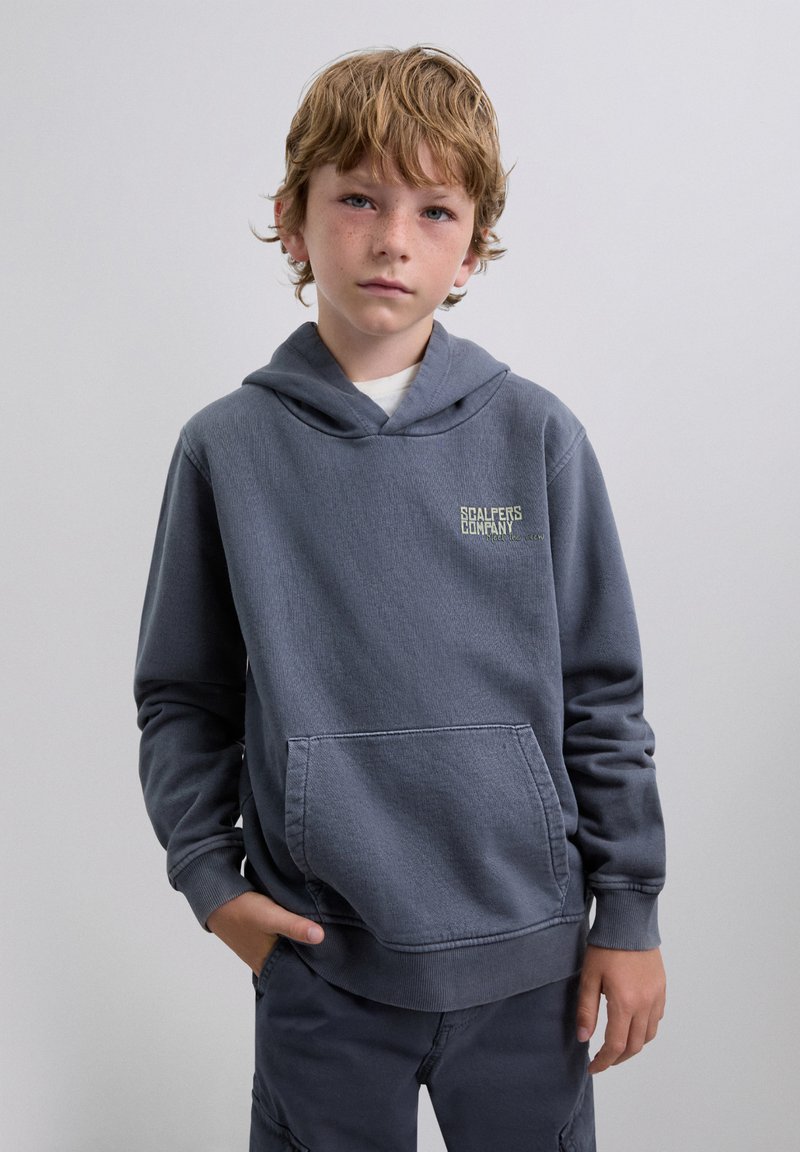 Sudadera gris con un bolsillo delantero, tela texturizada y un logotipo en el pecho. El modelo lleva pantalones oscuros a juego y tiene el cabello corto y despeinado.