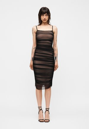 Γυναίκα που φοράει μαύρο διαφανές midi φόρεμα με ρυτίδες, λεπτές τιράντες και μαύρα ψηλοτάκουνα σανδάλια με λουράκια, στέκεται σε απλό φόντο.