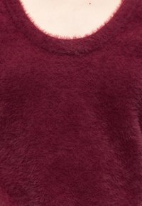 Felnőtt közeli képe, aki egy bolyhos, mély bordó pulóvert visel kerek nyakkivágással, látható a nyakának és a felső mellkasának egy része.