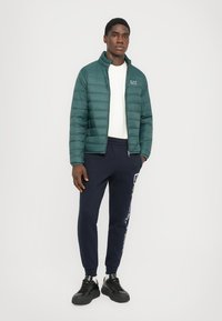 Grønn dunjakke med glidelås, kombinert med marineblå joggebukser med hvite grafiske detaljer. Modellen har på seg svarte joggesko.