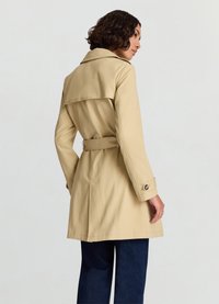 Trench-coat beige avec taille ceinturée, grand col et manches retroussées. Texture lisse avec un design mi-long et une fente discrète au dos.