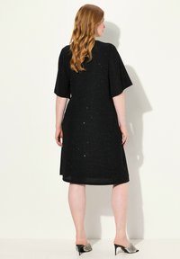 Robe noire midi en tissu métallique avec des manches courtes, présentant un subtil éclat. Associée à des mules à talons argentées.