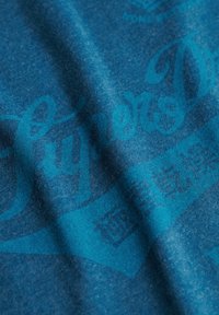 Camiseta de algodón azul con una textura suave, presenta un gran texto impreso en azul más oscuro. El diseño incluye letras estilizadas y patrones sutiles.