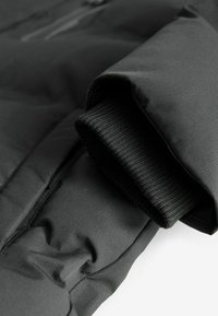 Manche de veste d'hiver noire avec poignet en maille côtelée visible et tissu extérieur lisse, vue en gros plan sous un angle.