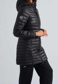 Svart puffjacka med huva, med dragkedja fram och två sidofickor. Designen inkluderar horisontella quiltade linjer och en logotypplåster.