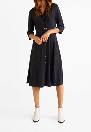 Femme portant une robe noire boutonnée jusqu'aux genoux avec des manches trois-quarts et une ceinture, associée à des bottines blanches à la cheville.