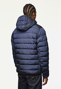 Marineblå pufferjakke med hætte, der har vandrette quiltede sektioner og en glat tekstur. Bæres med mørk denimjeans.