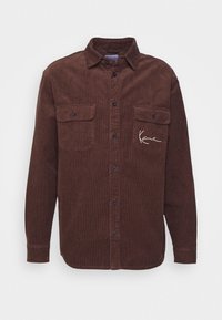 Braunes Cordhemd mit langen Ärmeln, zwei Brusttaschen, Knopfverschluss und einer weißen, gestickten Unterschrift auf der linken Tasche.