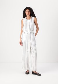 Ermeløs jumpsuit i hvit med blå striper, med knappelukking foran, belted midje og vide bukser. Svarte flate sko.