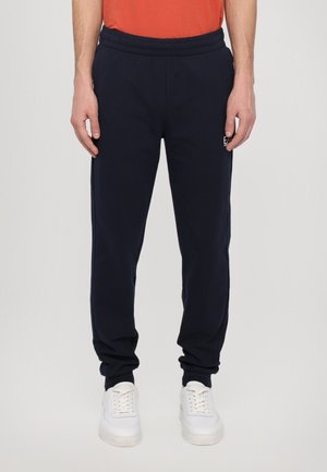 Mørkeblå joggebukser med elastisk midje, sidelommer og logo på låret, sammen med hvite sneakers.