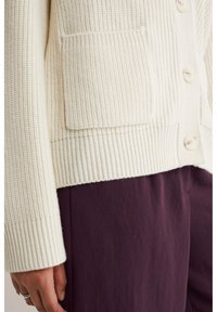 Cardigan lavorato a maglia di colore crema con texture a coste, chiusura con bottoni e una tasca frontale. Abbinato a pantaloni viola scuro.