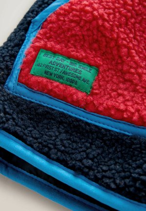 Sarkana un tumši zila flīsa auduma izstrādājums ar zilu apdari. Iespējama zaļa etiķete ar tekstu un logotipu. Strukturēta virsma ar mīkstu, pūkainu apdari.