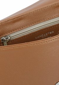 Borsa in pelle marrone con una chiusura zip in argento; presenta il nome del marchio "Lancaster" impresso su una fodera beige.