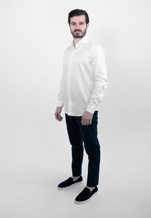 Witte lange mouw knoopshirt, donkere broek en zwarte instapschoenen. Glad stof, getailleerde pasvorm en klassiek kraagontwerp.