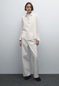 Wit linnen oversized overhemd met lange mouwen, gecombineerd met een broek met hoge taille en wijde pijpen, beide in een licht crèmekleurige tint.