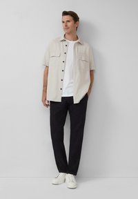 Korte mouwen beige shirt met donkere knopen, een witte T-shirt eronder, zwarte broek en witte sneakers, staande tegen een effen achtergrond.