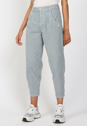 Jasnoniebieskie skrócone spodnie sztruksowe z przednimi plisami, noszone z białymi trampkami na stojącej osobie.