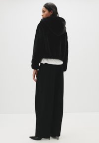 Črna krzna jakna s kapuco in skrajšanim krojem, ki ima mehak teksturo, elastičen rob, nošena čez belo srajco in v kombinaciji s širokimi črnimi hlačami.