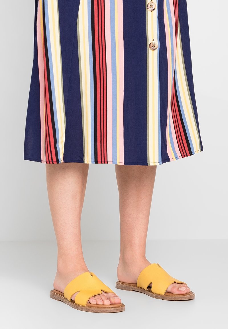 Sandales jaunes à glissière avec un design ajouré sur une semelle beige, assorties à une robe multicolore à rayures marines comportant des couleurs comme le rouge, le rose et le bleu.