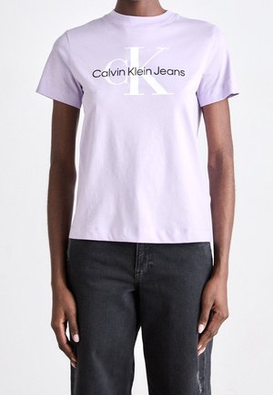Henkilö, jolla on päällään vaalean violetti Calvin Klein Jeans -t-paita ja mustat farkut, seisoo yksiväristä valkoista taustaa vasten.