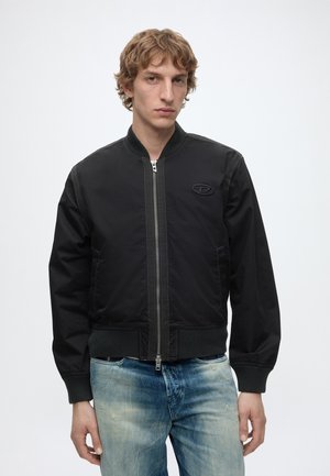 Giovane uomo con capelli ricci castano chiaro che indossa una giacca bomber nera con cerniera e jeans blu sbiaditi, in piedi davanti a uno sfondo bianco semplice.