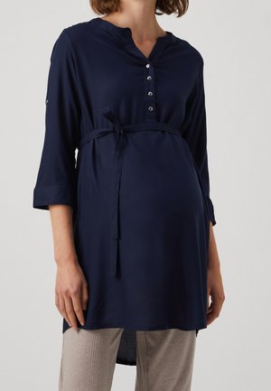 Gravid person iført en lang navy blå graviditetstunika med knaplukning ved halsen og taljebånd, kombineret med lyse beige bukser.